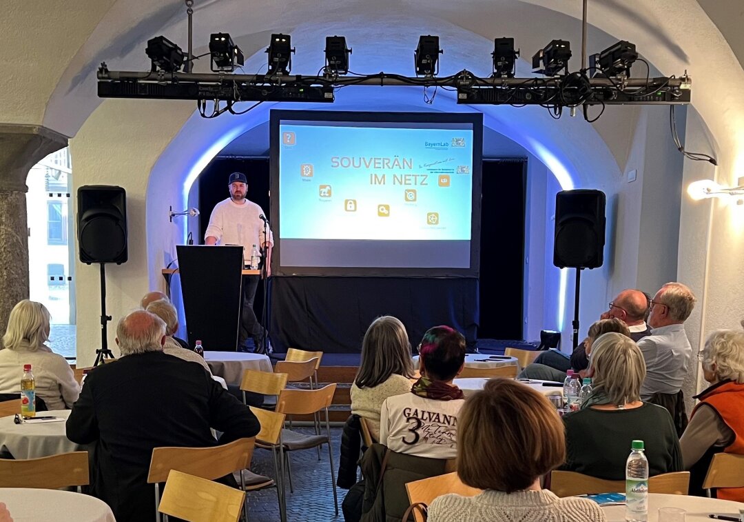 Lableiter Christopher Trengert steht vor einer Leinwand in einem Raum mit Gewölbedecke. An kleinen Tischen sitzen Kursbesucher mit Getränken.